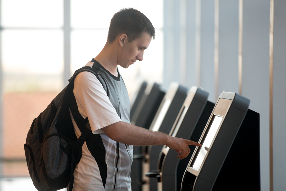 Young man with backpack touching interactive display at self-service transfer machine, doing self-check-in for flight or buying airplane tickets at automatic device in modern airport terminal building